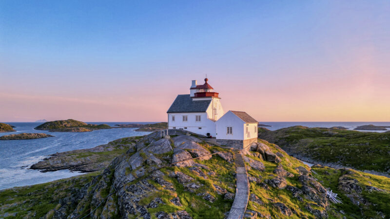 Et hvitt fyrhus med rødt tak står på en liten, steinete øy omgitt av hav. En smal tresti leder opp til bygningen. Landskapet badet i varmt lys fra solnedgangen, og små øyer ligger spredt i bakgrunnen.