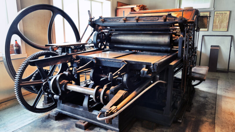 En stor, gammel trykkpresse i et museumsmiljø. Maskinen er laget av mørkt metall med store tannhjul, valser og et svinghjul på venstre side. Den står i et lyst rom med vinduer bak og trykkerirelaterte gjenstander på veggene.