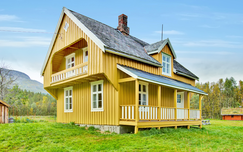 Slette med gress og et stort, gult, gammelt hus. Åser og fjell i bakgrunnen, samt blå himmel. None mindre bygninger i bakgrunnen til venstre og høyre.