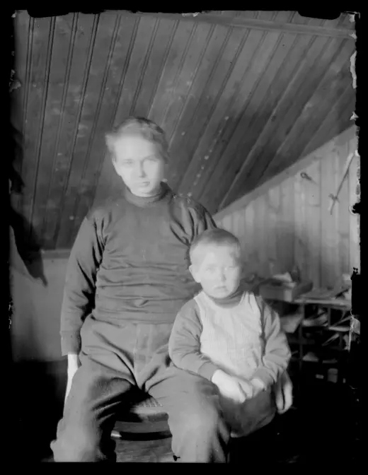 Et gammelt svart‑hvitt foto av to barn som sitter tett sammen på en stol i et rom med skråtak og trepanel på veggene. Det minste barnet sitter foran, mens det andre sitter litt høyere bak. I bakgrunnen står enkelte møbler og gjenstander delvis i skygge.