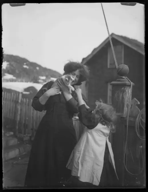 Et eldre svart‑hvitt fotografi som viser to personer utendørs. En voksen person står nær en bygning av tre, mens et barn står foran og rekker armene opp mot den voksne. I bakgrunnen er det et gjerde, et fjellparti med snø og ulike detaljer fra et gårds- eller kystmiljø.