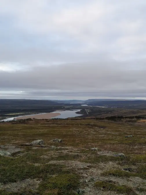 «Utsikt over et vidstrakt landskap med lave åser og en lang, smal vannflate som slynger seg gjennom terrenget. Forgrunnen består av lav vegetasjon i dempede grønn‑ og bruntoner. I bakgrunnen ligger fjell og et bredt, grått skydekke som gir scenen et dempet og kjølig preg.»