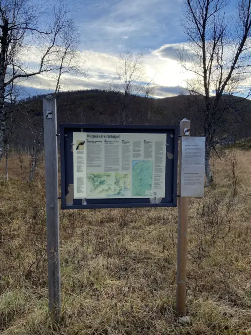 Informasjonsskilt i høstlandskap med noen åser i bakgrunnen, noen trær rundt og blå himmel med noen skyer.