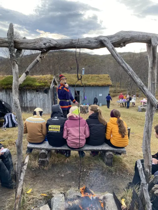 Et bål foran i bildet og noen barn som sitter med ryggen til . Foran dem en man i samisk kofte som snakker i mikrofon. Flere gamle hus i bakgrunnen og en ås bakerst i bildet. Lettskyet himmel.