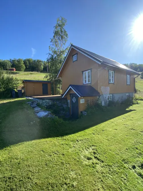 Et okergult hus på en grønn plen. Bak huset en gresslette i bakke og så en skog bak der.