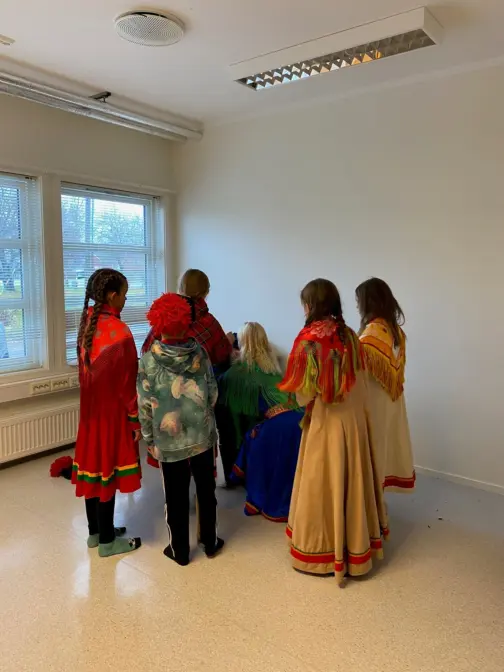 Barn kledd i samiske kofter står med ryggen til et rom