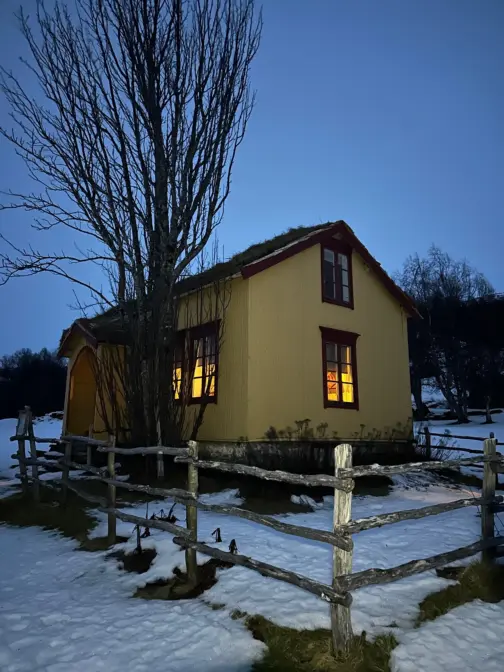 Gammelt okerfarget hus i grålysningen. Snø på bakken. Et stort tre foran huset. Et gjerne går rundt huset.