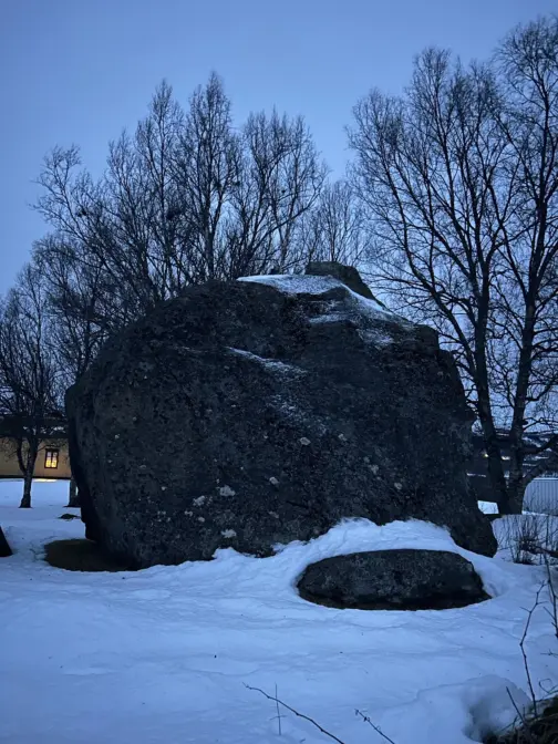 En stor stein i grålysningen. Snø på bakken. Store tre bak steinen.